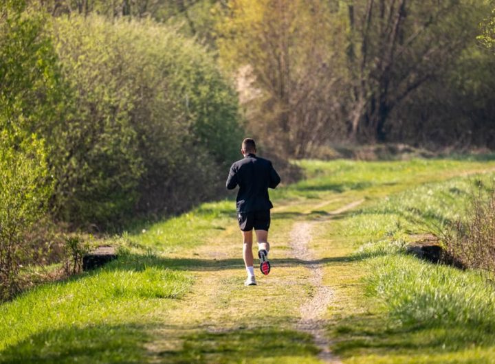 A man jogs on a scenic, grassy forest path surrounded by lush greenery under daylight.