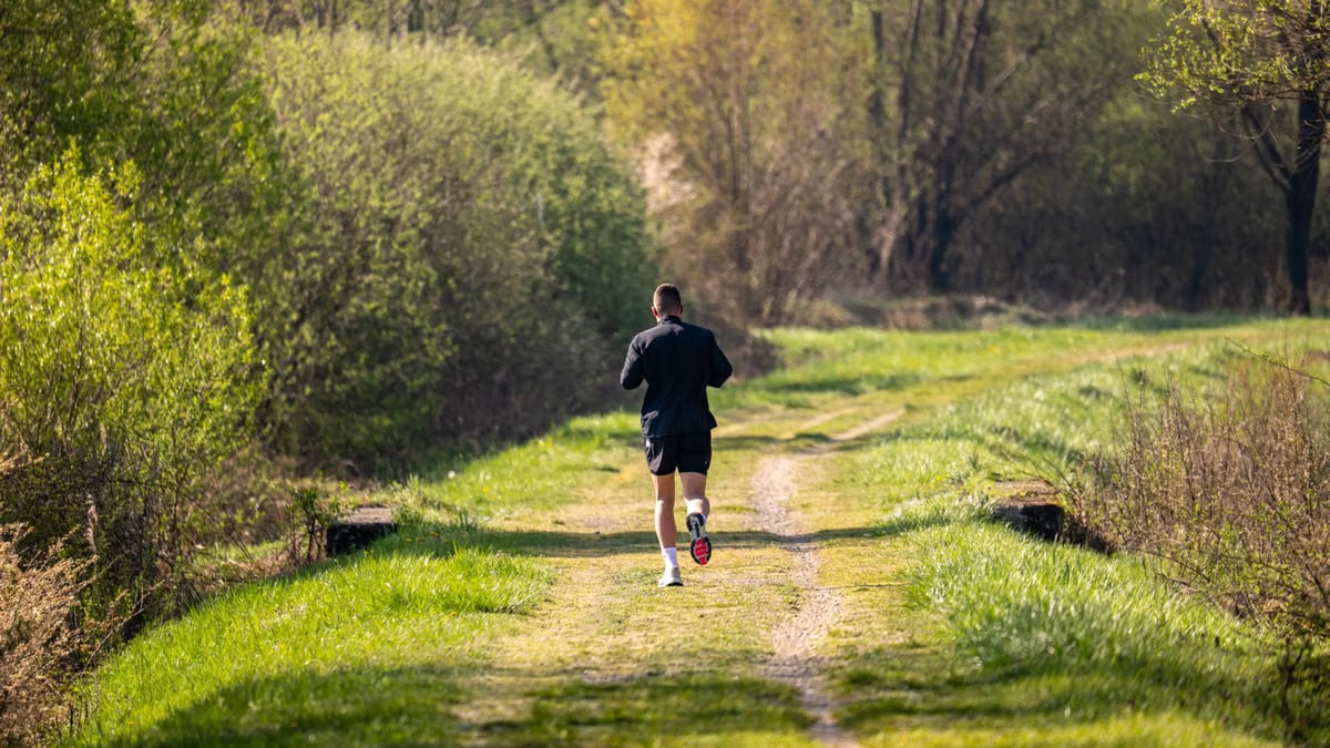 A man jogs on a scenic, grassy forest path surrounded by lush greenery under daylight.